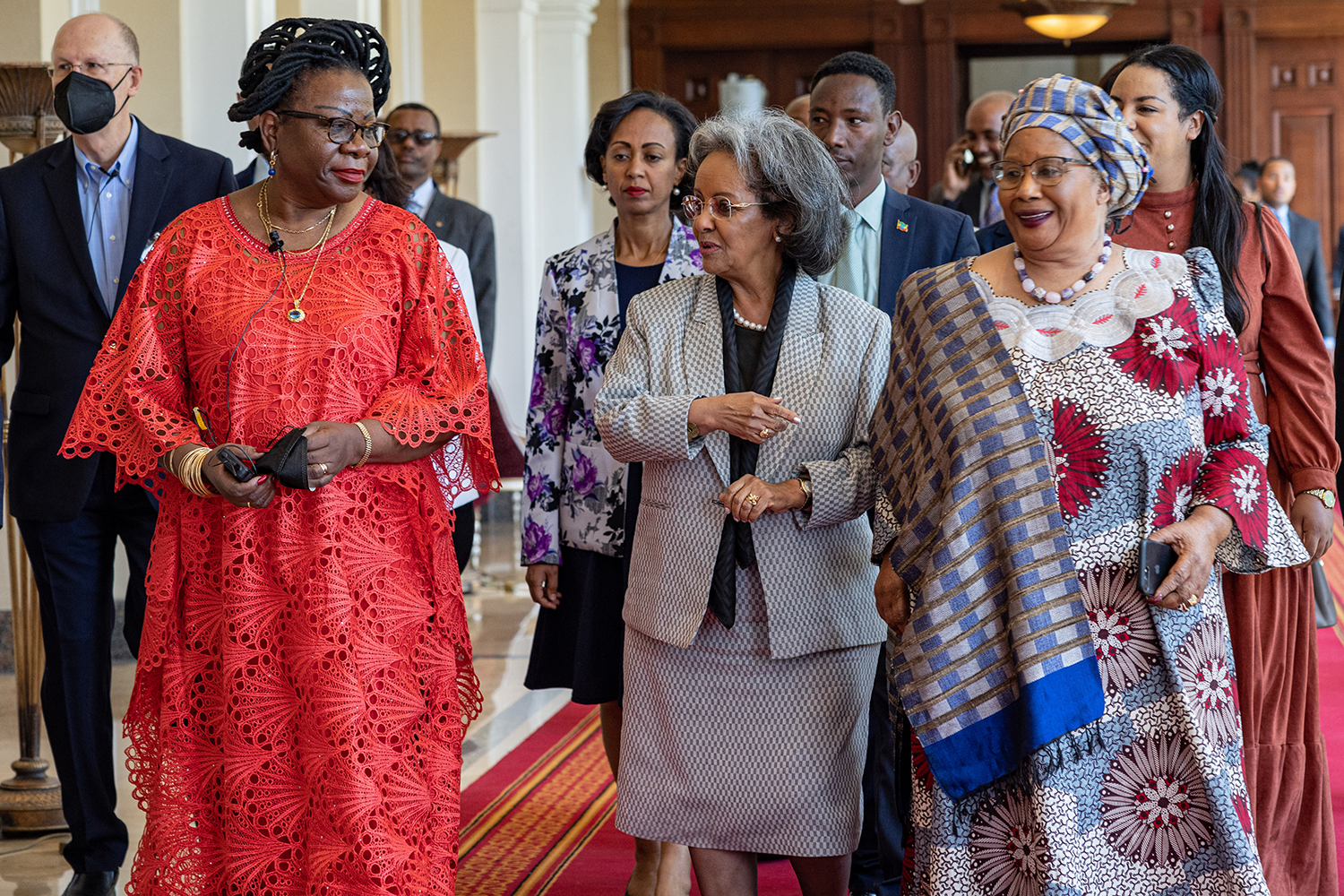 The Rt. Hon. Luisa Diogo, former Prime Minister of Mozambique, in the left of the picture and Her Excellency Joyce Banda, former President of Malawi, in the right of the picture welcome Ethiopian President, Her Excellency Sahle-Work Zewde, in the middle of the picture to the 2022 Roundtable meeting in Addis Ababa. The three women converse while walking down a corridor to the meeting, accompanied by a group of Ministers and security personnel.
